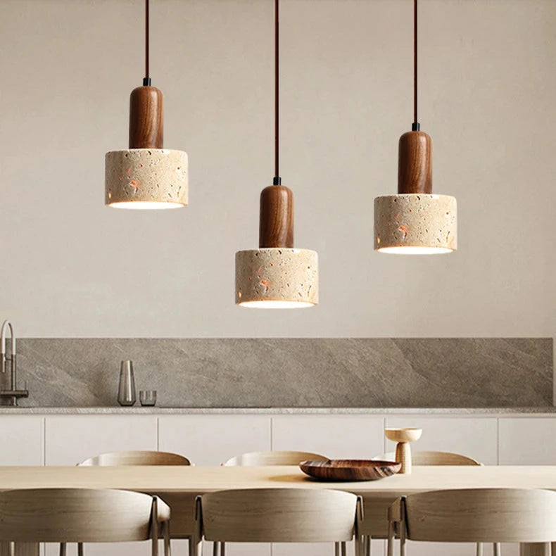 A minimalist kitchen features two Morsale.com Natural Travertine Pendant Lamps with wooden tops and stone-like concrete shades, illuminated by LED bulbs, hanging above a light wood table with a shallow wooden bowl. The background includes a marble backsplash and a counter with minimal décor.