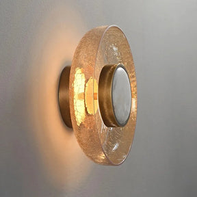 The Morsale Art Deco Cracked Glass Wall Sconce showcases a modern design with a crackled glass shade that emits a warm, glowing light. Its circular form includes a central metallic detail, enhancing the elegant ambiance against a grey wall.