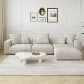 A modern living room showcases a Morsale Corduroy Modern Sectional Couch Sofa, highlighted by premium corduroy fabric and cushions. A uniquely shaped white coffee table sits on a light rug, framed art decorates the walls, and neutral-toned curtains enhance the light wood flooring.