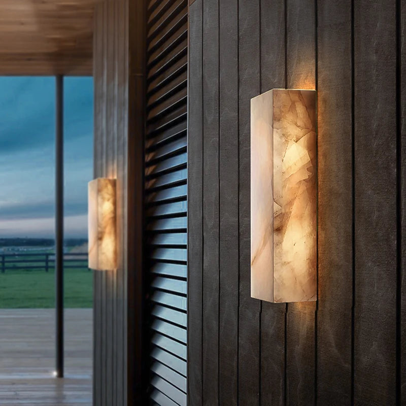 Two Morsale Outdoor Marble LED Wall Lamps (IP65 waterproof) emit warm light on a dark wood-paneled wall, illuminating a modern patio with views of fields in the background.