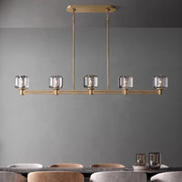 The Morsale Ventoux Linear Crystal & Brass Chandelier with five crystal shades hangs above a dark dining table with beige and gray velvet chairs against a gray wall, accented by two glass candle holders on the table.