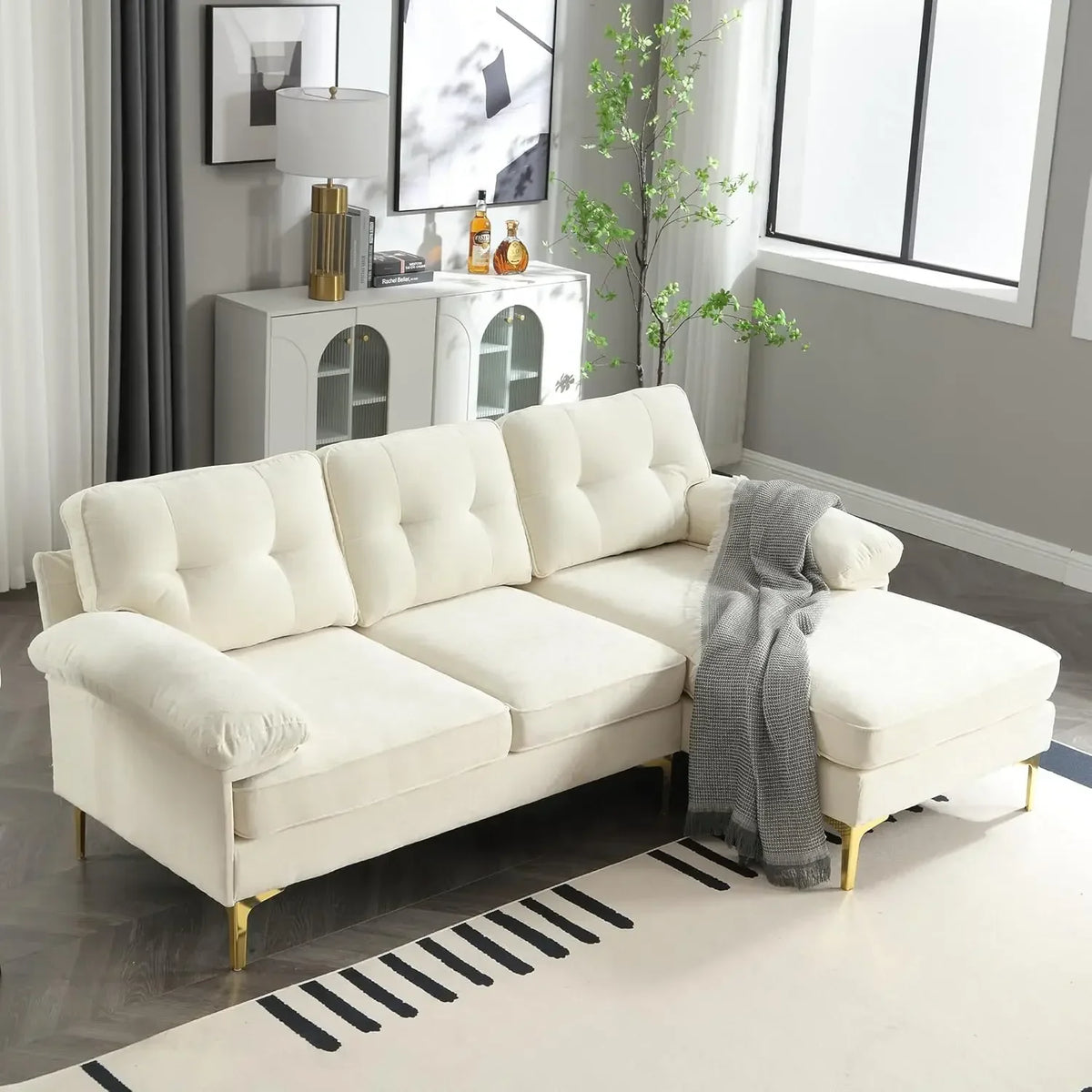 A modern living room showcases an 83" Morsale sectional sofa with a chaise in luxury chenille fabric adorned with a gray throw. Behind it, a white cabinet holds decorative items. Nearby are a tall lamp and potted plant, while a large window floods the space with natural light.