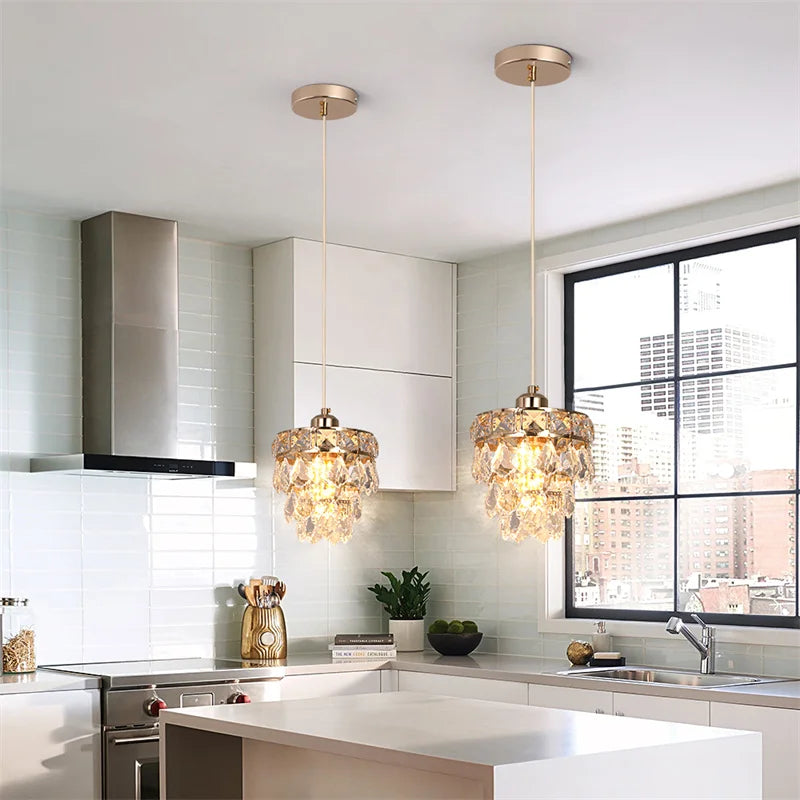 A modern kitchen with white Lazzo cabinets, a countertop, and backsplash. Two Morsale.com Lazzo Kitchen Table Pendant Light Fixtures with handmade crystals adorn the ceiling. A large window offers a view of city buildings. A range hood and sink are also visible.