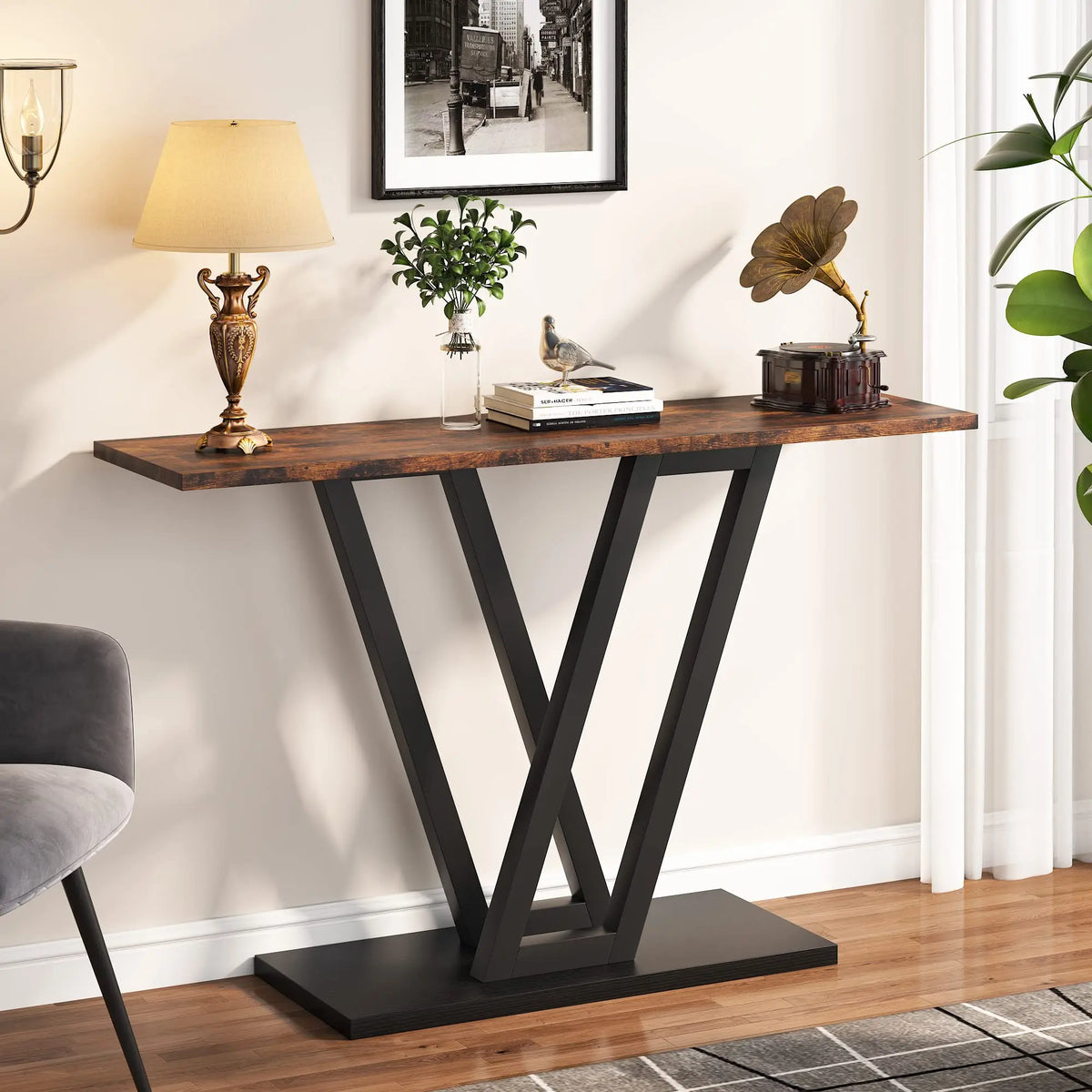The Bradford Entryway Table by Morsale showcases a contemporary V-shaped black metal base and wooden top, complemented by decor items like a lamp, books, a metal sculpture, greenery, and artwork on a light-colored wall—ideal for the living room.