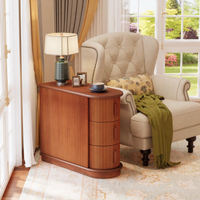 Elegant room with a beige armchair, a green blanket, and floral cushion. A Morsale Galway Solid Wood End Table with three drawers holds a table lamp, cup, and eyeglasses. Yellow curtains frame a large window offering a garden view.