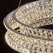The Bacci Modern Crystal Chandelier by Morsale is an elegant circular piece adorned with intricate diamond-shaped crystals. Illuminated with LED bulbs, it casts a warm glow, while its metal frame adds modern flair against a dark background.