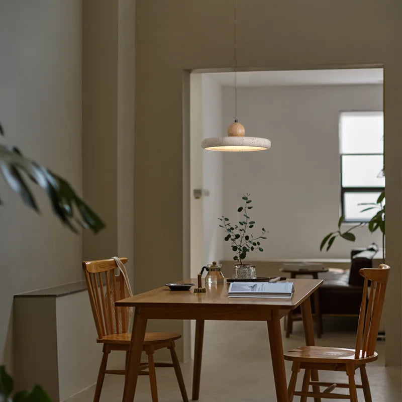 A minimalist dining area featuring a small table with dark legs and a light wooden top, surrounded by four chairs. The beige walls are adorned with a black-and-white abstract framed picture. Overhead, modern Natural Travertine Kitchen Pendant Light Fixtures from Morsale.com fitted with LED bulbs illuminate the space, which is subtly decorated with small items.