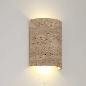 A Natural Travertine Art Deco Wall Lamp from Morsale.com, crafted from beige, porous stone and shaped into a cylinder, is mounted on a white wall. The light radiates softly from both the top and bottom, generating an ambient glow around the fixture that accentuates its ART DECO design.