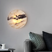 A cozy living room showcases the Morsale Moonshade Natural Marble Sconce Light with warm LED lighting. Below, a gray couch with textured pillows sits beside a small side table holding a white cup against a light gray wall.