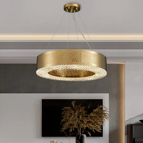 A contemporary interior features the sleek Alleri Hammered Gold & Crystal Chandelier by Morsale, adorned with LED bulbs and a textured finish, elegantly suspended by wires. Below, a television is mounted on a neutrally-toned wall, while a vase filled with dried plants enhances the sophistication of an adjacent table.