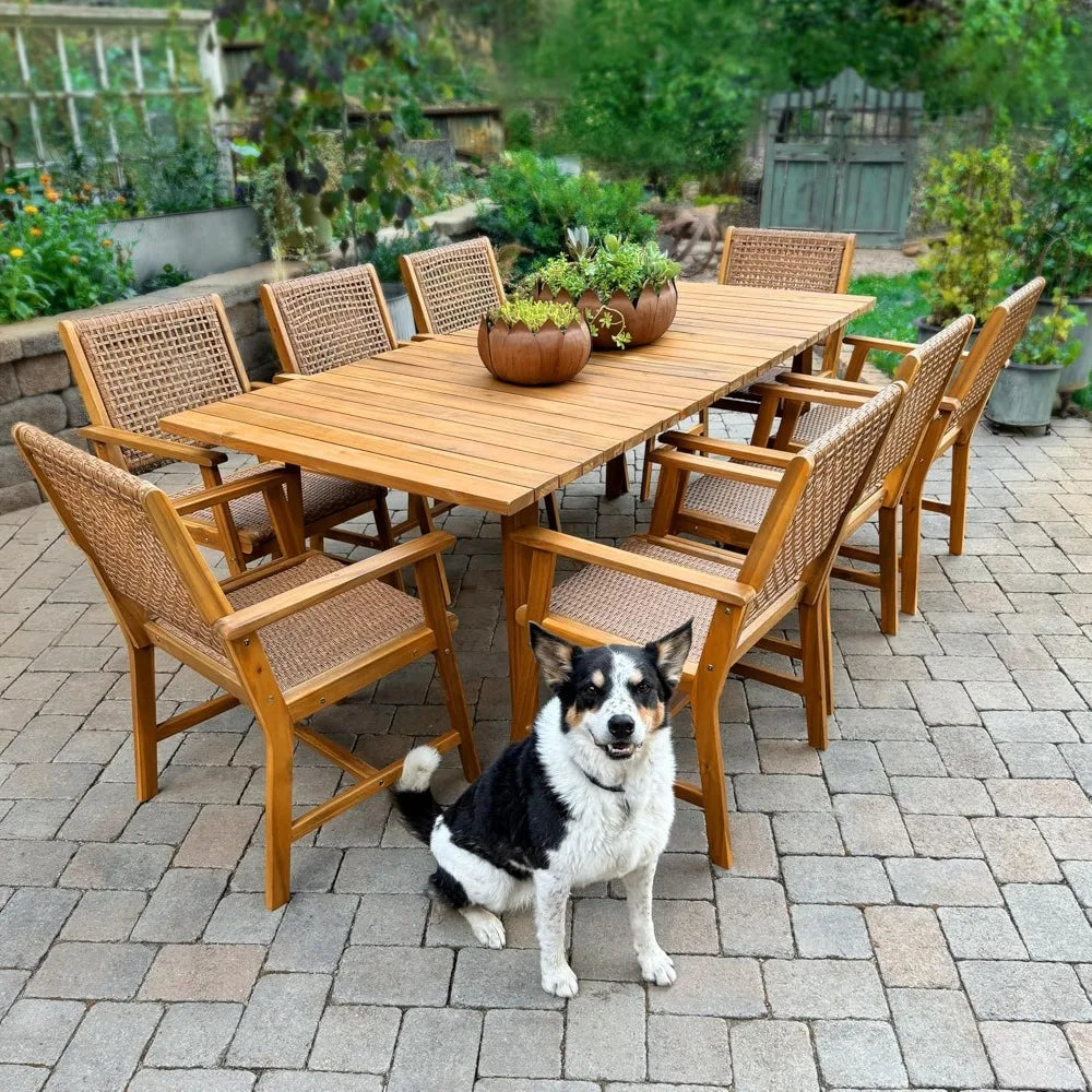 A dog sits by the Morsale Brackley 9-Piece Outdoor Dining Set on a brick patio, where the teak table features three succulent-filled pots and is set against lush tropical greenery and a wooden gate backdrop.