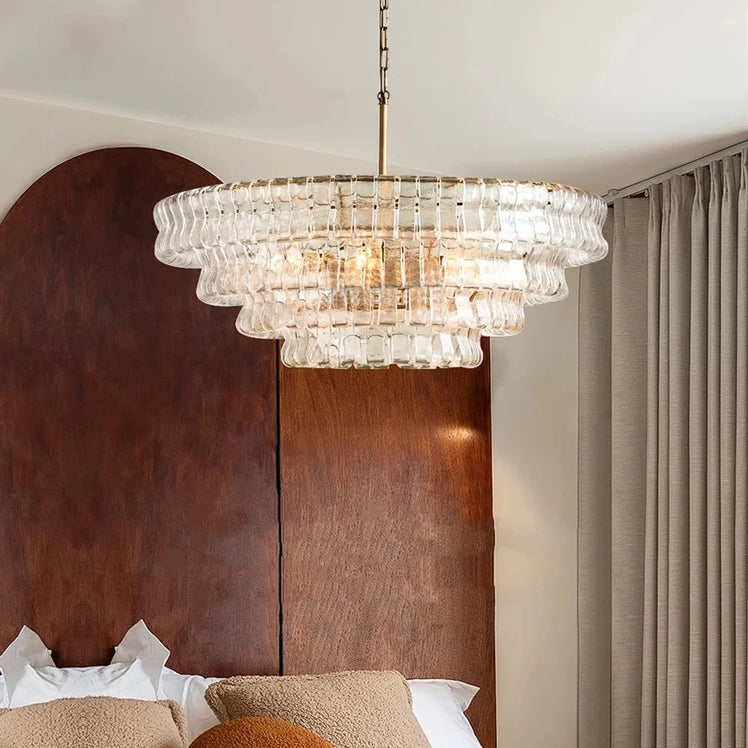 The Morsale Riviera Contemporary Chandelier hangs above a bed with white pillows, brown cushions, a curved wooden headboard, and beige curtains, bringing modern elegance to the room.