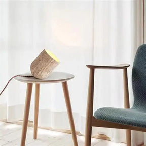 A small, cylindrical Modern Travertine Table Lamp from Morsale.com, with a concrete texture, tilts at an angle on a light-colored surface. Emitting a warm yellow light reminiscent of natural yellow cave stone, it casts a soft glow on the wall behind it. The power cord is visible trailing off to the side.