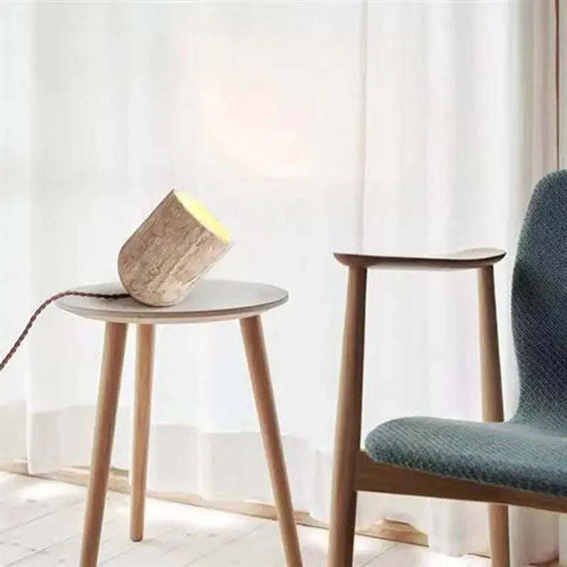 A small, cylindrical Modern Travertine Table Lamp from Morsale.com, with a concrete texture, tilts at an angle on a light-colored surface. Emitting a warm yellow light reminiscent of natural yellow cave stone, it casts a soft glow on the wall behind it. The power cord is visible trailing off to the side.