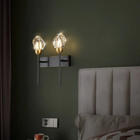 A contemporary Diamante Crystal Wall Sconce by Morsale, featuring two crystal-like bulb coverings, is mounted on an olive green wall above a beige upholstered headboard. Made with copper material, the LED lamp illuminates the area. A mug rests on the bedside table beneath which a power outlet is located below the sconce.