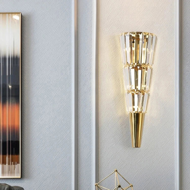 A modern interior showcases the Gio Stainless Steel & Crystal Sconce by Morsale, casting LED light on a textured white wall. Below, a geometric gold sculpture rests on a round black side table adjacent to a dark gray couch. On the left, a blurred abstract painting enhances the chic ambiance created by these contemporary wall lamps.