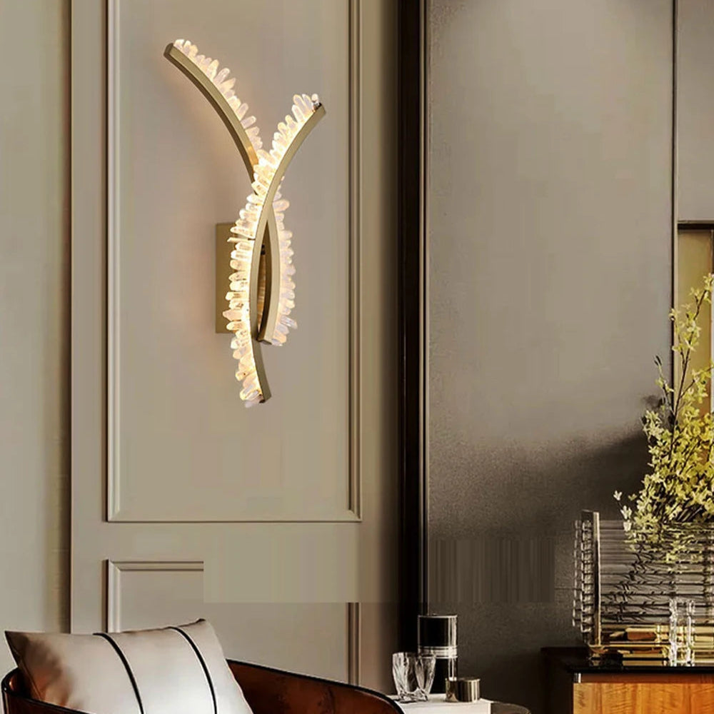 A stylish interior showcases the Orion Crystal Wall Sconce by Morsale, casting a warm glow with its curved design. Below, an elegant chair with a patterned cushion complements a small table adorned with glassware and flowers, enhancing the modern style lighting.