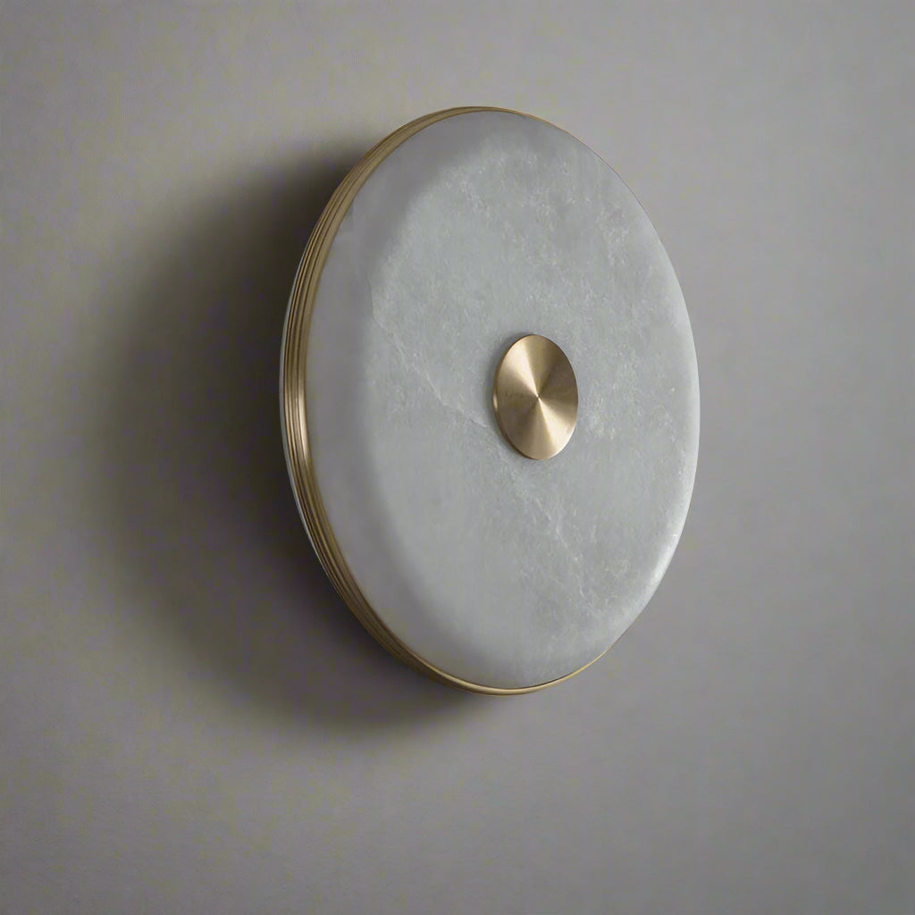 The Moonshade Natural Marble LED Wall Light by Morsale features a frosted glass surface and central brass accent, epitomizing modern design while mounted on a dark, textured wall.