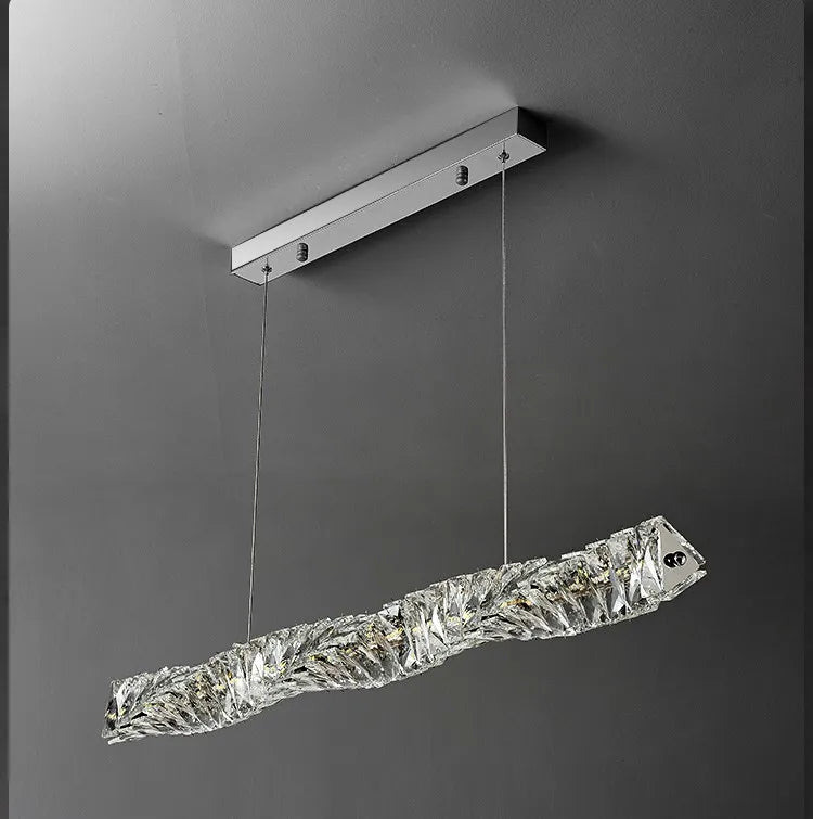 The Bacci Modern Crystal Dining Room Light Fixture by Morsale.com features a sophisticated rectangular pendant design with a gold ceiling mount and two thin wires suspending a crystalline, textured, elongated LED fixture. The light emits a warm glow, enhancing the elegant ambiance against a neutral gray backdrop—an ideal choice for contemporary dining spaces.