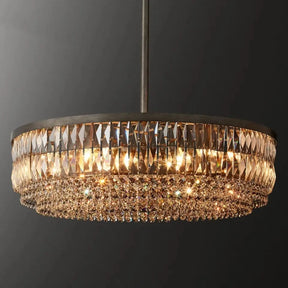 A luxurious, circular Bacci Crystal Chandelier by Morsale.com hangs from the ceiling against a dark background. The chandelier features multiple rows of K9 crystal prisms that create an elegant, shimmering effect when illuminated.