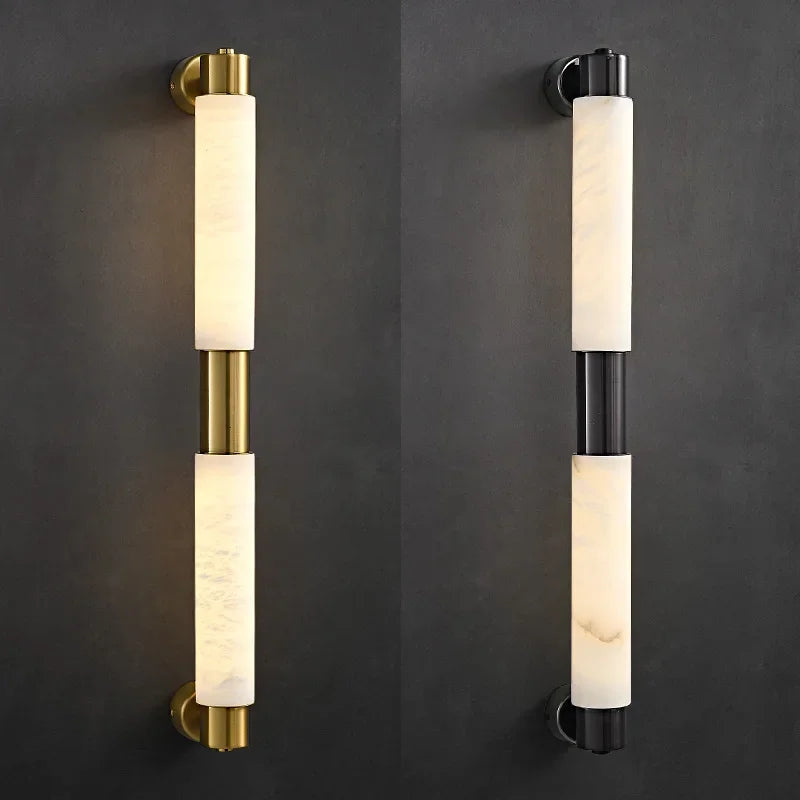 Mounted on a dark wall are two wall sconces: the left sconce showcases a sleek design with a brass finish, while the right one is an elegant Morsale Moonshade Natural Marble Wall Lamp in matte black. Both feature cylindrical cream-colored shades that offer a diffused light effect.