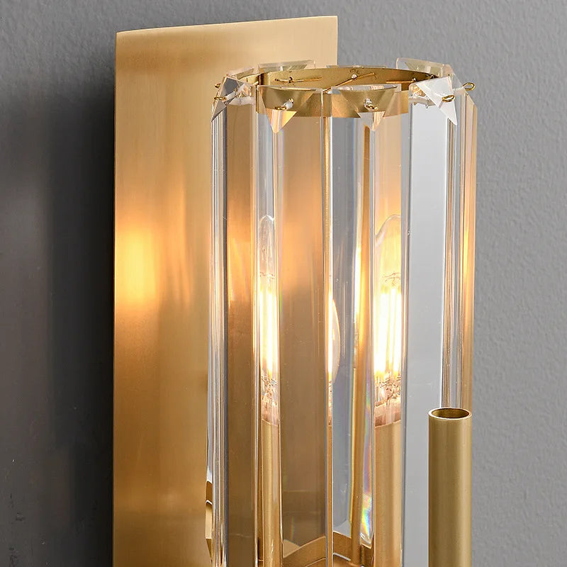 Introducing the Gio Double Head Crystal Wall Sconce from Morsale, a modern lighting fixture featuring a gold rectangular base and vertical glass prisms that encase two LED bulbs. This exquisite piece casts a warm glow, enhancing an elegant and sophisticated ambiance when placed against a gray wall.