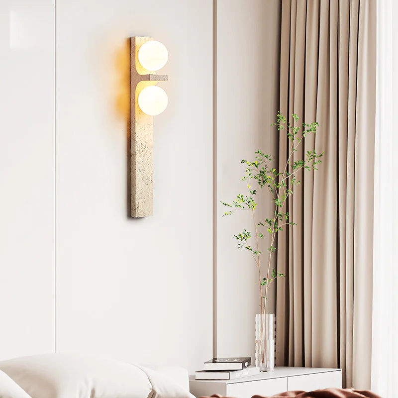 A modern Japan Style wall-mounted light fixture from Morsale.com, featuring two glowing circular LED bulbs, is vertically installed on a light-colored wall. Below the Natural Travertine Modern Wall Lights, to the left, a green leafy plant is partially visible.