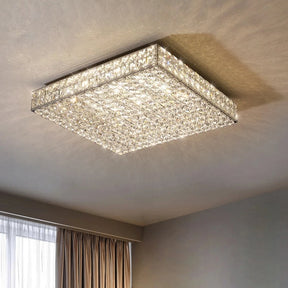 A Morsale.com Bacci Clear Crystal Light Fixture is mounted on the ceiling, illuminating the room with a warm, elegant glow. The luxurious light fixture has a shimmering surface due to numerous clear crystals. A large window with beige curtains is partially visible in the background.