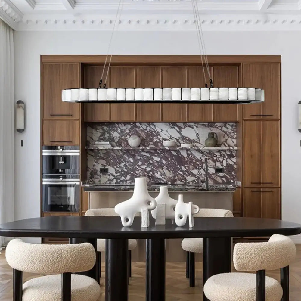 
                  
                    Modern dining room featuring a black oval table, abstract white vases, beige textured chairs, wooden cabinetry, marble backsplash, and the Hampton Marble Dining Room Chandelier by Morsale with dimmable LED lighting and white shades above.
                  
                