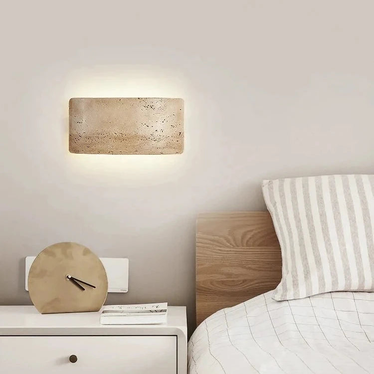 The Natural Travertine LED Wall Light Fixture by Morsale.com is an interior wall sconce featuring a rectangular, curved stone-like cover that emits a warm, soft glow. This energy-efficient LED lighting creates a halo effect on the white wall, emphasizing the texture and natural imperfections of the stone surface.