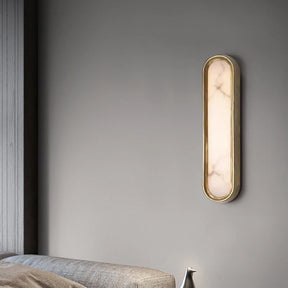 The Moonshade Natural Marble LED Wall Lamp by Morsale, featuring a sleek, oval-shaped design with a brass frame and a diffused light resembling natural marble, is mounted on a gray wall. Below it, part of a gray upholstered bed or sofa can be seen, highlighting modern style and minimalist interior design.