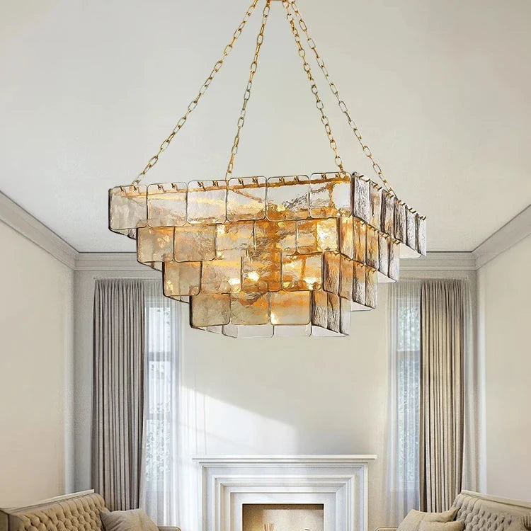 A living room with two beige sofas facing each other, centered around a white coffee table adorned with candles. Overhead hangs the Morsale Welle Glass Chandelier, showcasing its modern design with polished steel and square glass panels illuminated by LED bulbs. In the background, a decorative fireplace and tall curtains complete the setting.