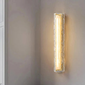 The Morsale Postmodern Luxury Glass & Copper Wall Sconce, with its contemporary rectangular design and textured glass cover, casts a warm glow against a light gray wall, cultivating a cozy and elegant atmosphere.