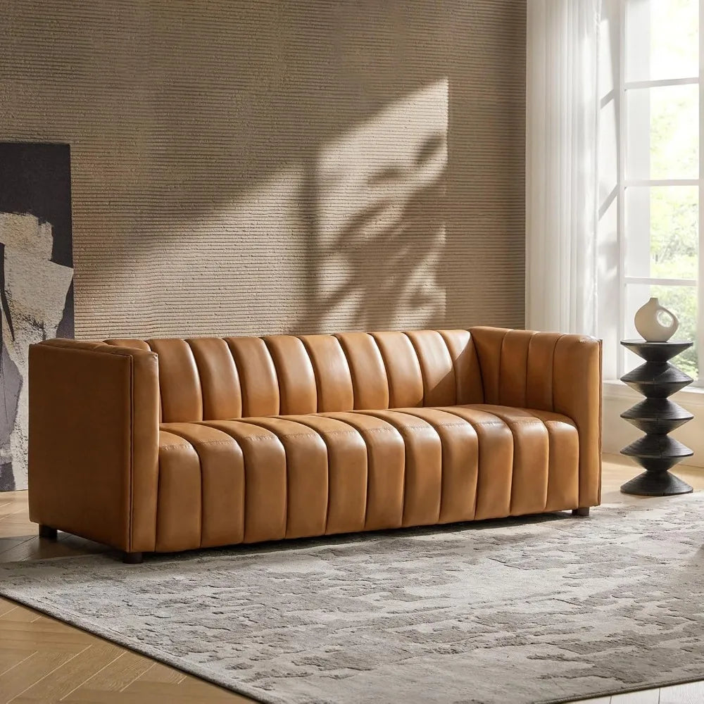A modern living room showcases a Morsale Biagio 83" Genuine Leather Sofa with vertical tufting, complemented by a textured beige wall. A contemporary black sculptural side table displays decor, while sunlight floods through a large window, accentuating a patterned gray rug.