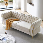 The bright living room showcases Morsale's luxurious 87” Living Room Sofa Couch in cream, featuring button tufting and gold accents. A mustard throw adorns one arm. The space includes velvet furniture, soft lighting, plush rugs, and a view of greenery through sheer curtains.