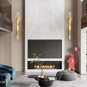 A modern living room showcases a tall white marble fireplace adorned with sleek Gio Crystal Sconces by Morsale. Positioned in front of the fireplace, a black coffee table topped with a plant adds charm. The decor is completed by blue chairs and a red bear sculpture holding a balloon.