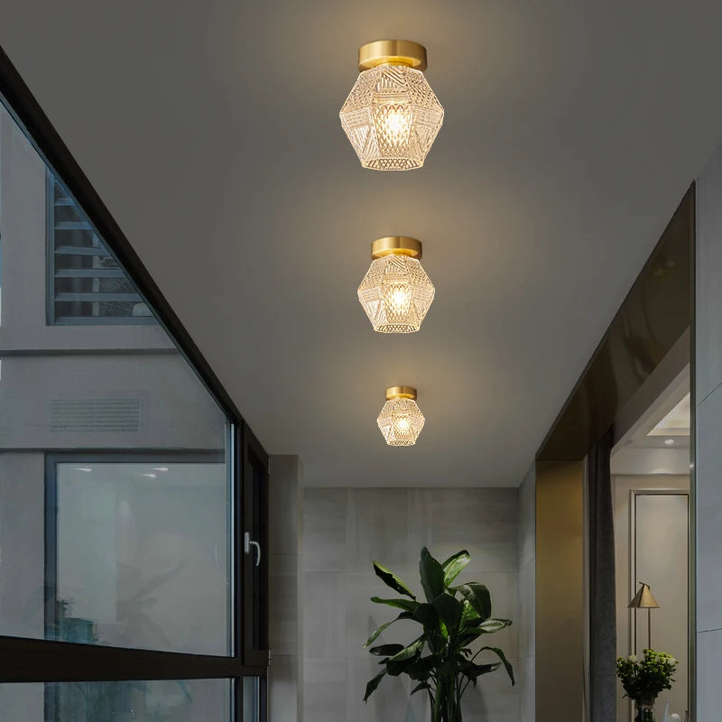 The hallway exudes a modern vibe with three Morsale Palermo Ceiling Lights, each showcasing geometric glass shades and brass fittings, enhanced by energy-efficient LED bulbs. A large window on the left bathes the area in natural light, while a green potted plant introduces a hint of nature below.