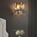 The Lazzo Crystal Wall Sconce by Morsale.com shines warmly against a dark, elegant wall with vertical grooves. Nearby are a beige upholstered headboard and a small table topped with a white flower arrangement.