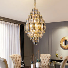 A luxurious dining room showcases the Lazzo Crystal Modern Chandelier by Morsale, now fitted with LED bulbs, hanging above the table. Tufted chairs flank the space, complemented by a round mirror and decorative items on a side table. A curtain elegantly drapes over a large window on the left.