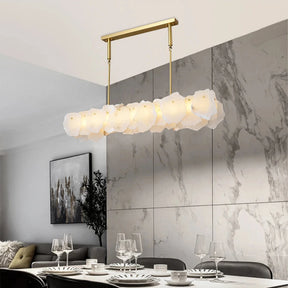 A modern dining area featuring the Natural Marble Dining Room Chandelier by Morsale, a long, rectangular fixture with a hexagonal frosted glass design hanging over a white oval table adorned with a black bowl and clear vase. The space is surrounded by beige upholstered chairs and set against a minimalist tiled wall.