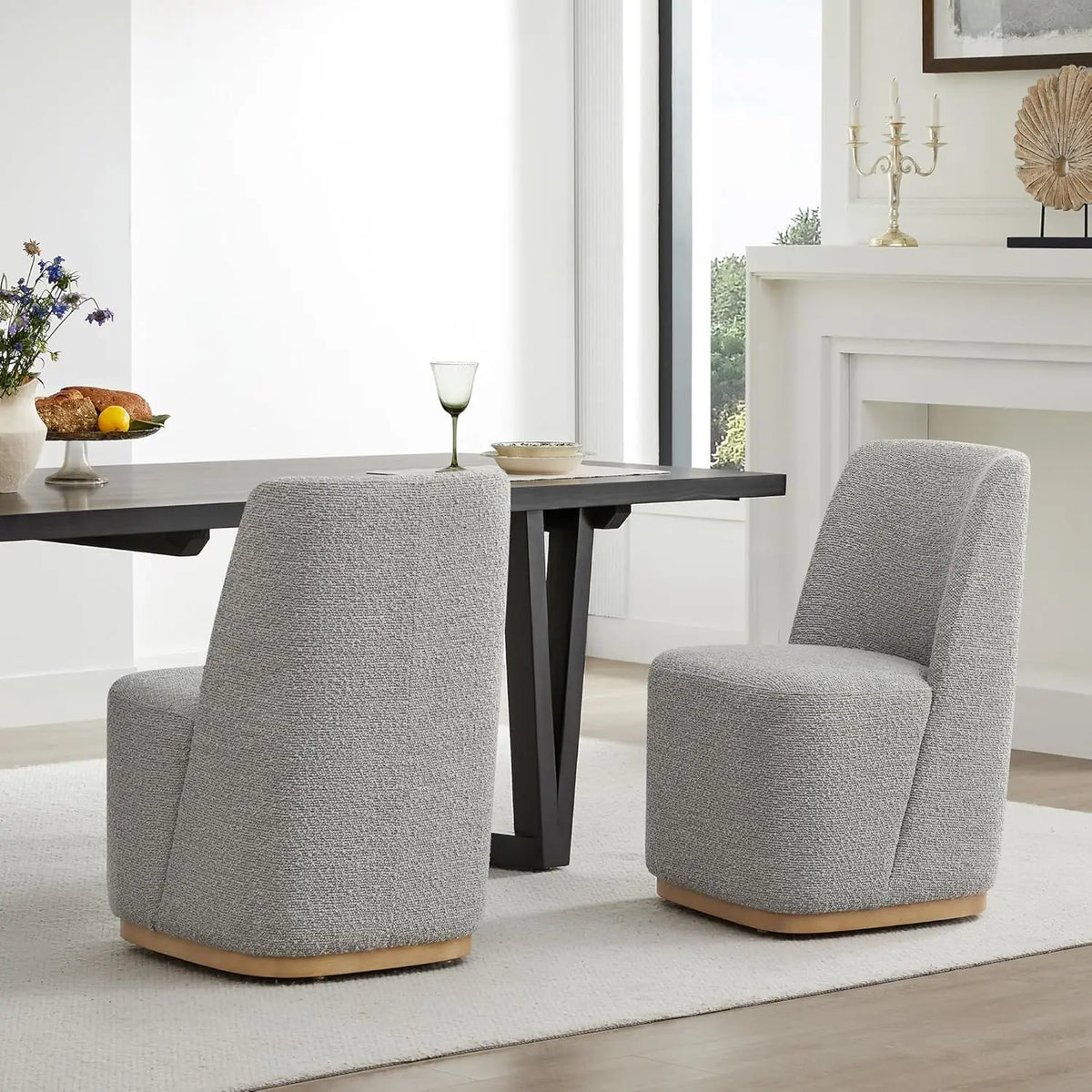 A modern dining area features a rectangular black table with two Morsale Boucle Dining Chairs With Casters in sleek gray. A glass filled with green liquid sits on the table, and a candleholder along with decorative items embellishes the white mantel. Large windows illuminate the space naturally.