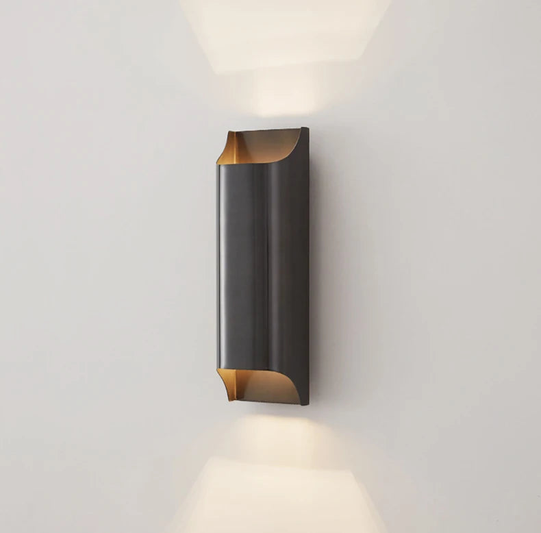 The Morsale Rame Copper Sconce is a contemporary cylindrical LED wall lamp with an open top and bottom that emits warm light. It boasts a sleek, dark metallic finish evocative of copper, and is mounted against a light-colored wall.