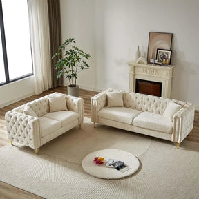 The living room showcases a Morsale 2-Piece Loveseat Sofa Set in cream chenille upholstery on a light rug, accompanied by a round coffee table with books and flowers. A potted plant sits by a large window, while framed art and candles grace the small fireplace, enhancing modern elegance.