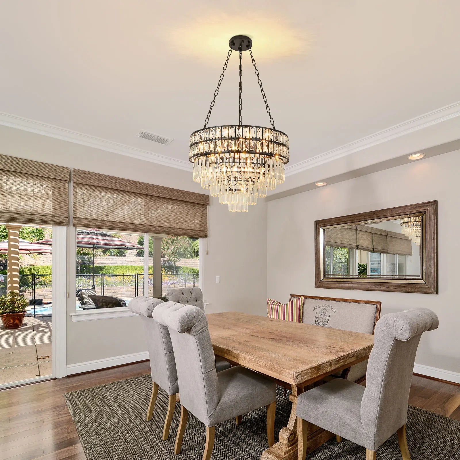 A dining room with a wooden table, six cushioned chairs, and the Gio Modern Crystal Chandelier from Morsale exudes modern style. A framed mirror graces the wall, while large windows reveal an outdoor pool area. The room features light-colored walls and hardwood flooring.