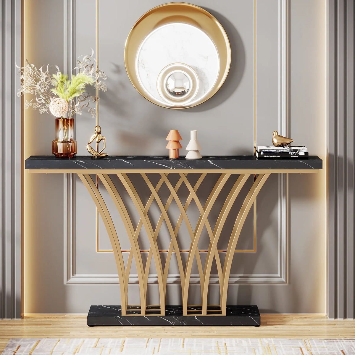 A sophisticated entryway features the Morsale Bassett Entryway Table with a black faux marble finish and geometric gold metal base, complemented by a gold circular mirror, dried floral arrangement, abstract statue, books, and bird figurine. Neutral walls enhance the elegant setting.