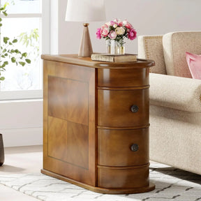 The Chester Solid Wood End Table With Drawers by Morsale, featuring three curved drawers inspired by Modern Morocco, is placed beside a beige sofa. A lamp, a book, and a vase of pink flowers adorn the top. Sunlight streams in through the window, partially illuminating a nearby plant.