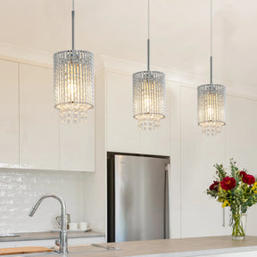 A modern kitchen showcases Morsale's Alleri Crystal Pendant Lights casting a glow over a countertop with a faucet. The space is adorned with white cabinets, a stainless steel refrigerator, and a vibrant bouquet of red and yellow flowers for contrast.