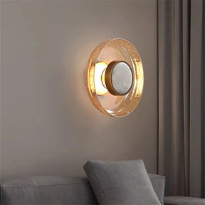 The Art Deco Cracked Glass Wall Sconce by Morsale boasts a modern design with a circular, translucent, amber-toned shade that emits a warm glow. Mounted on a neutral-colored wall next to a gray cushioned chair, it features an Art Deco touch. Its crackled glass element enhances the minimalist and cozy ambiance.