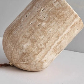 A small, cylindrical Modern Travertine Table Lamp from Morsale.com, with a concrete texture, tilts at an angle on a light-colored surface. Emitting a warm yellow light reminiscent of natural yellow cave stone, it casts a soft glow on the wall behind it. The power cord is visible trailing off to the side.