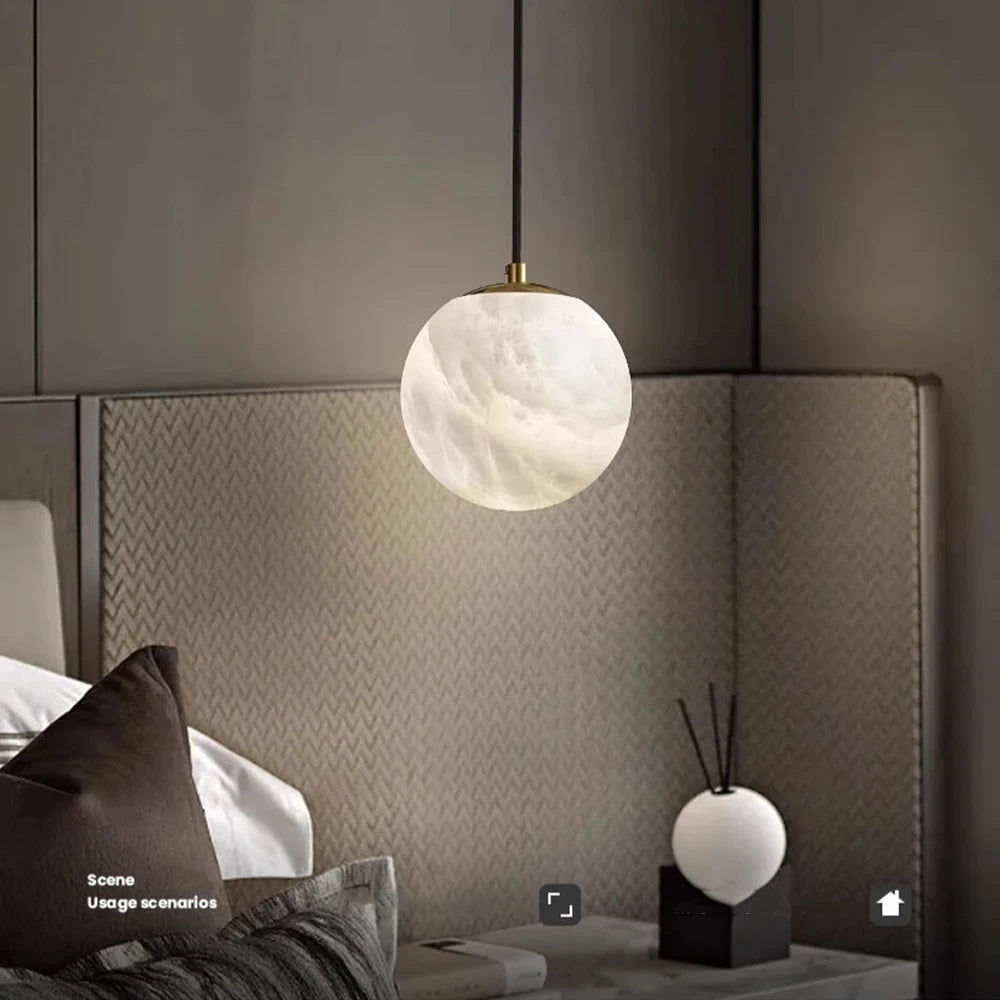 A cozy bedroom with a modern style showcases the Moonshade Natural Marble Ball Pendant Light by Morsale, featuring LED bulbs above the bed. The bed is adorned with gray and white pillows. A textured, chevron-patterned wall adds charm, while a small decorative sphere and a reed diffuser enhance the nightstand.